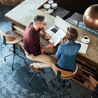 GettyImages-942622752 Two people seated at a bar having a conversation. One of them is holding a page with data tables on it while looking at a laptop screen.