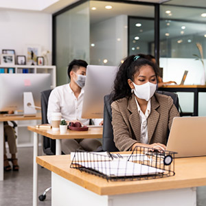 Masked coworkers seated at desks using computers Smiling client with arms crossed holding their right thumb up.