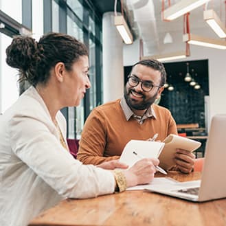 We'll set you up in five simple steps. Two people seated at a table while holding notepads and having a conversation in front of an open laptop.